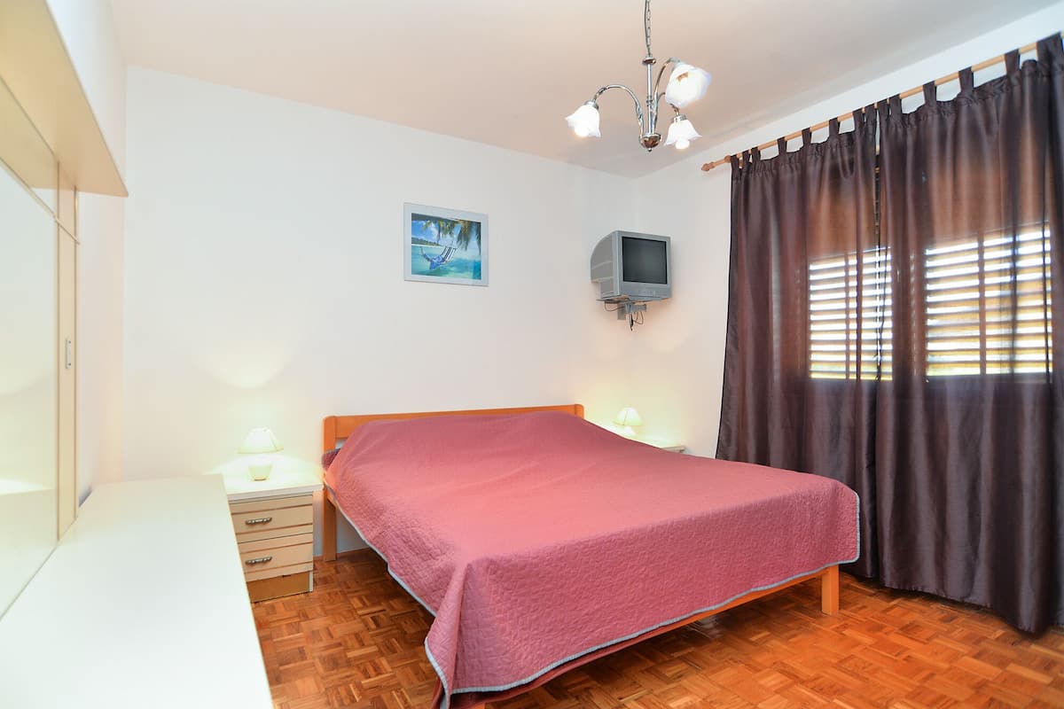 A bedroom with a wooden bed, a bedside table with a lamp, a TV mounted on the wall, and a window with curtains.