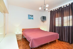 A bedroom with a wooden bed, a bedside table with a lamp, a TV mounted on the wall, and a window with curtains. - Apartment 1956 (Pula)
