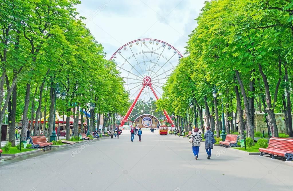 A park with a Ferris wheel, benches, and people walking.