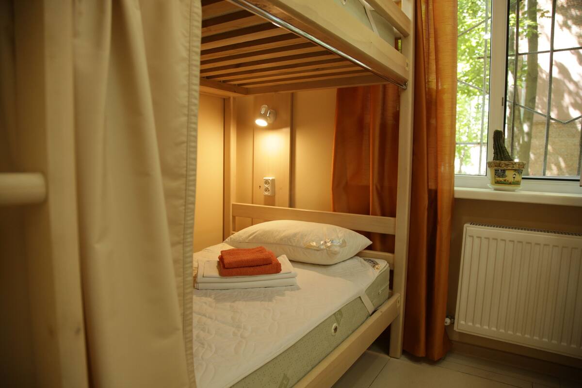 Un letto singolo con cuscino e asciugamani piegati, una lampada da comodino, una finestra con tende e un termosifone.