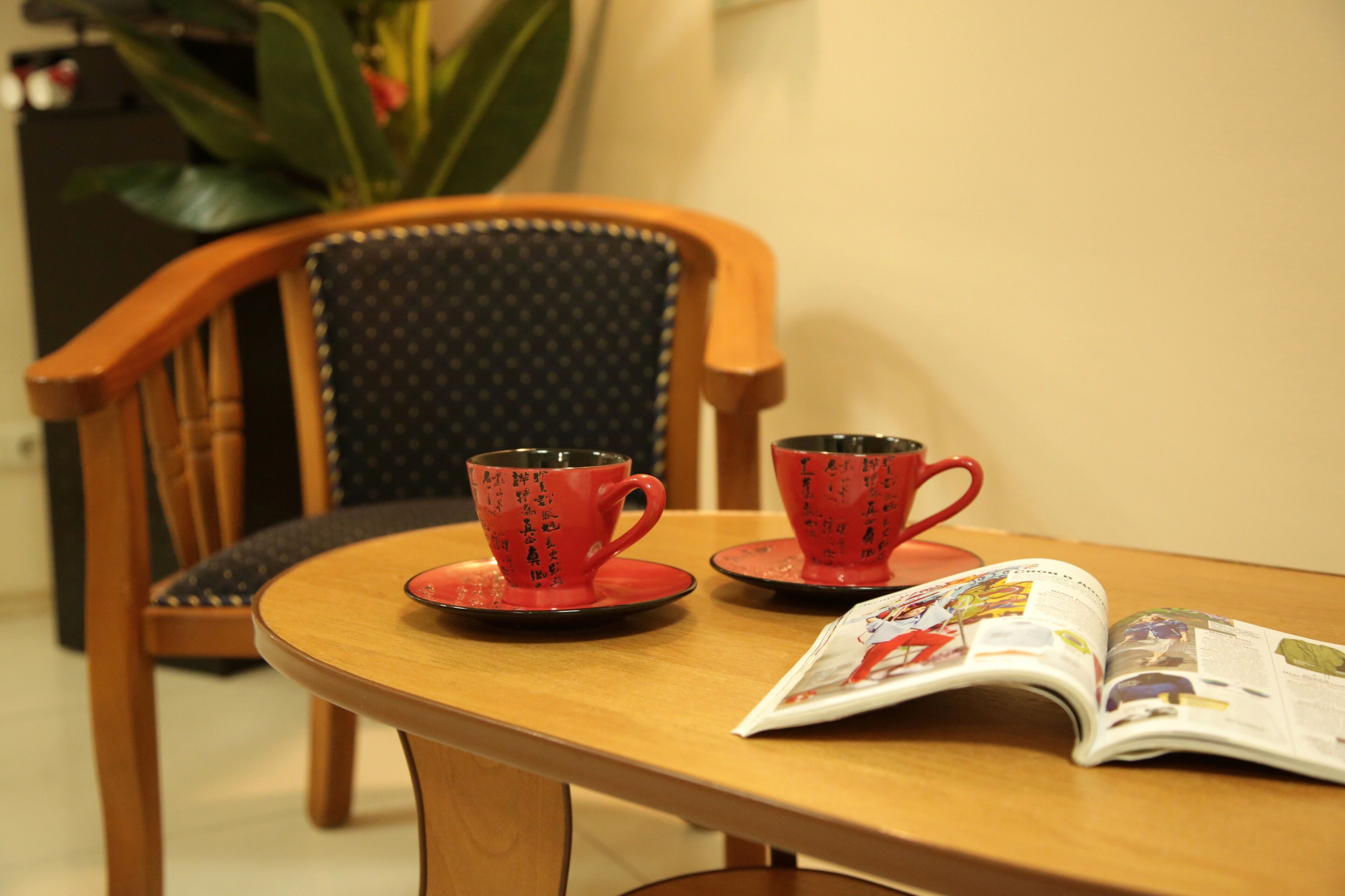 A wooden table with two red cups and saucers, a magazine, and a chair with a dark patterned cushion.