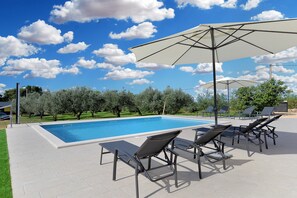 Una piscina con sillones de playa y sombrillas, rodeada de árboles y un cielo despejado.