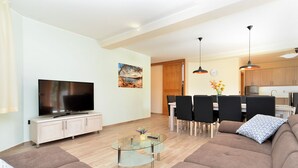 Un salon moderne avec un téléviseur à écran plat, une table basse en verre et un canapé. On y trouve également un coin repas avec une table et des chaises, ainsi qu’une cuisine à l’arrière-plan.