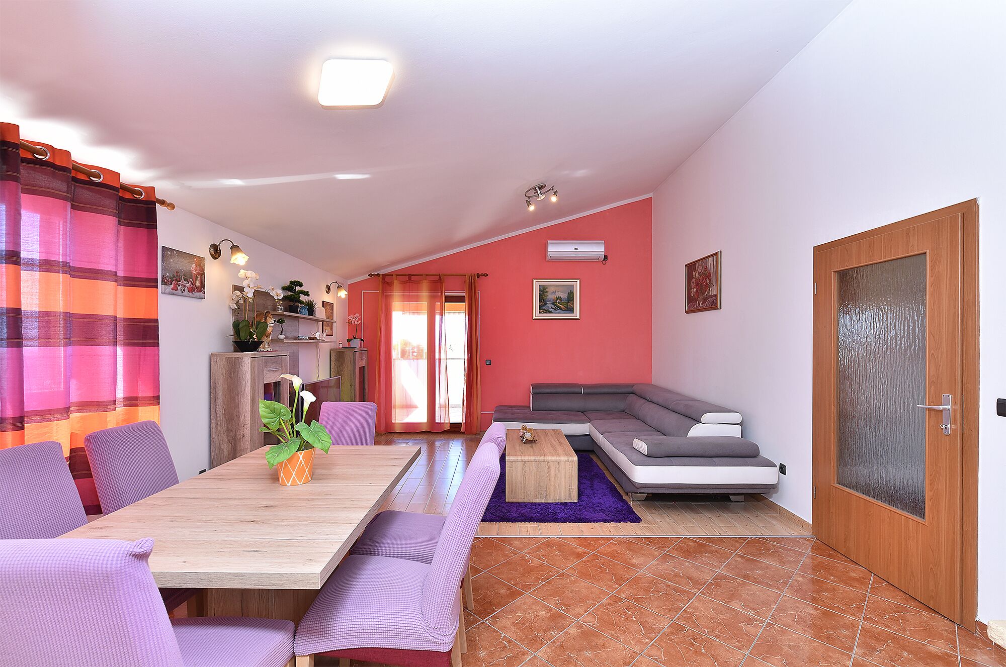 A living room with a wooden dining table, purple chairs, a grey sofa, and a red wall.