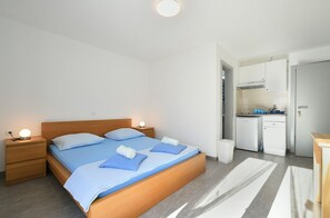 A modern bedroom with a wooden bed, two bedside tables with lamps, a small kitchenette, and a bathroom.