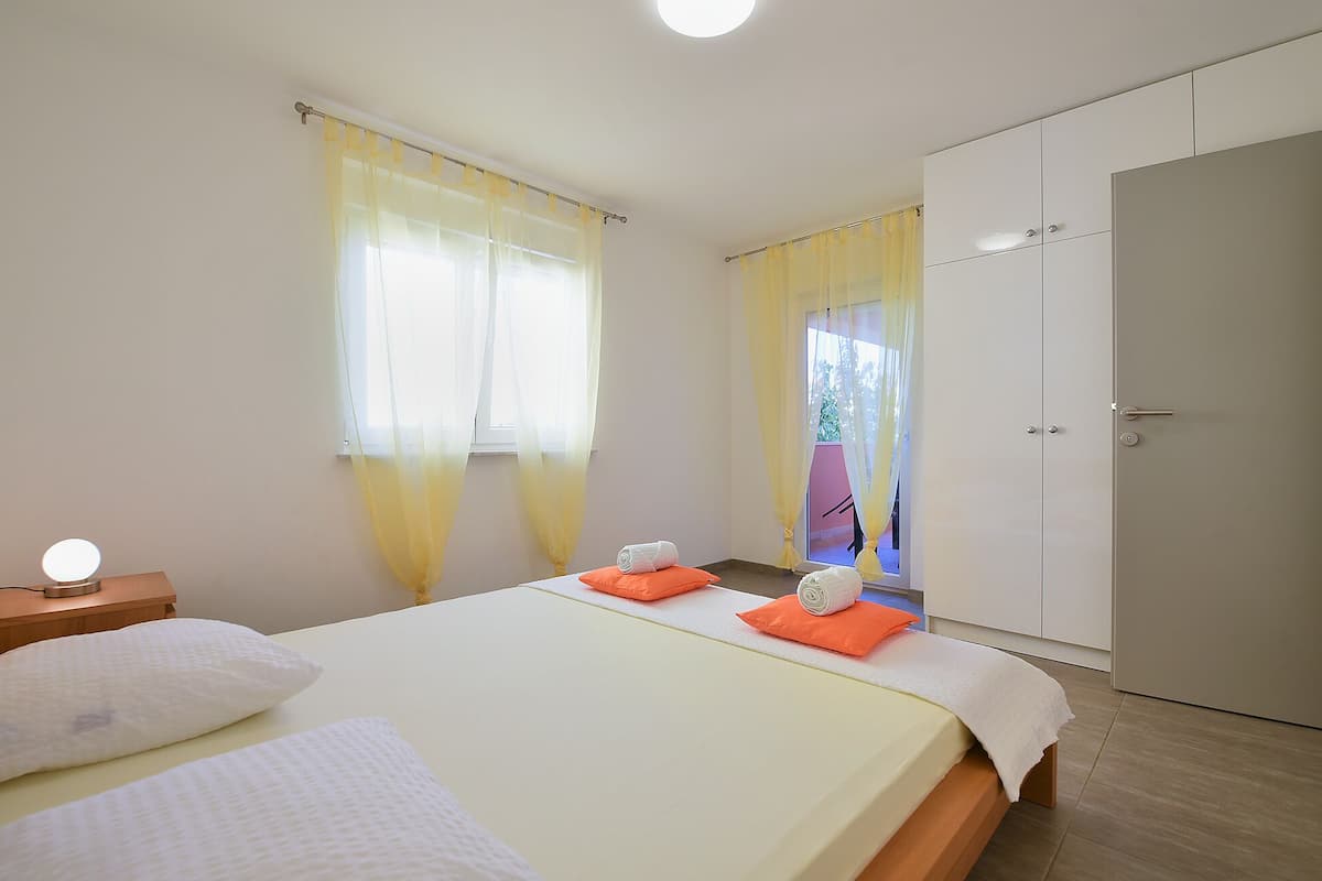 A bedroom with a bed, two pillows, a bedside table with a lamp, a wardrobe, and a window with curtains.