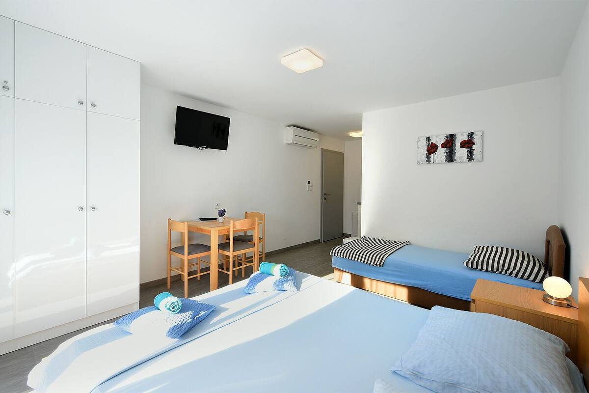 A bedroom with a bed, a desk, and a chair. There is a television on the wall and a door leading to another room.