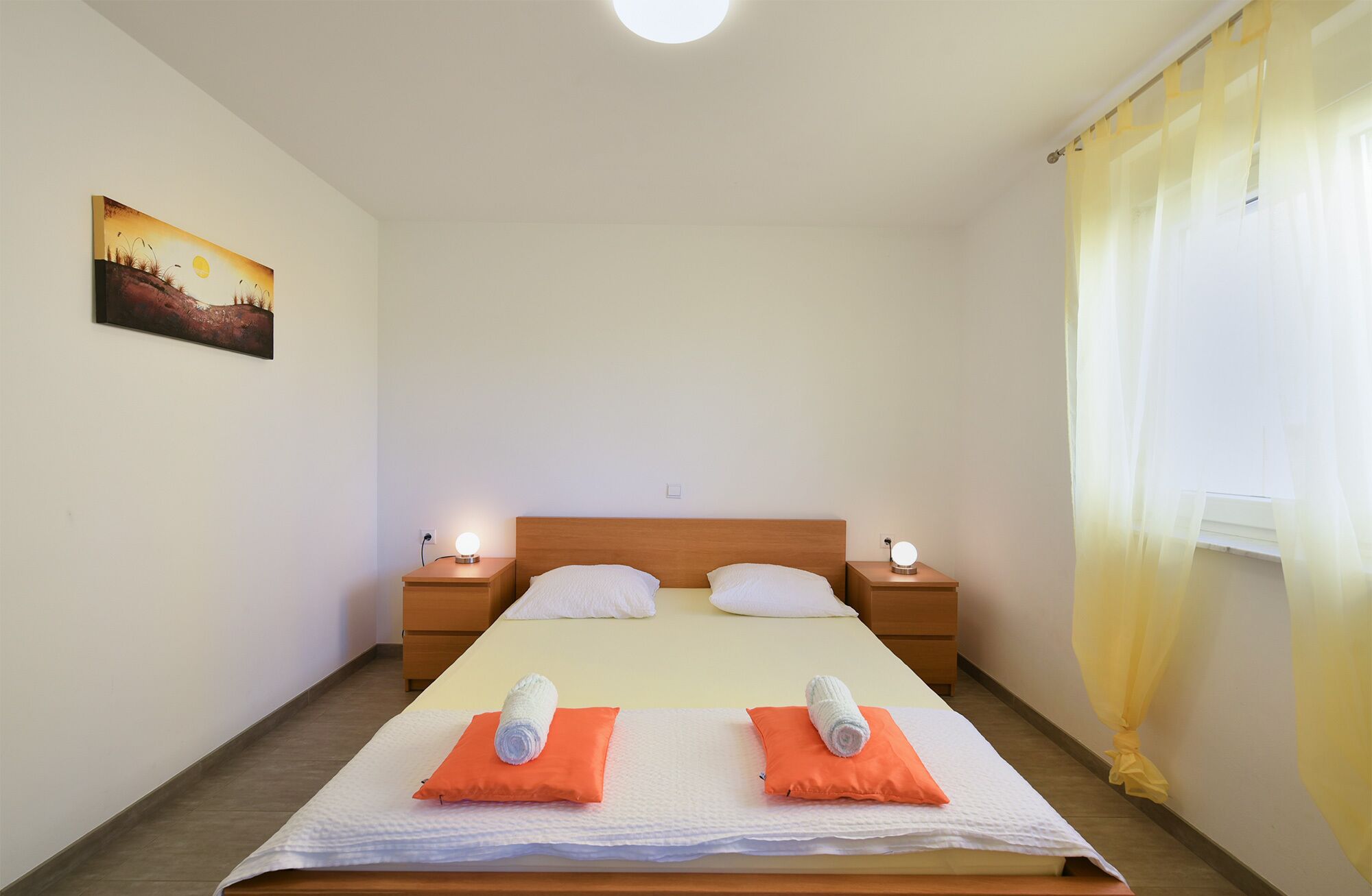 Una camera d'albergo con un letto, due comodini, un quadro appeso al muro e una finestra con tende.