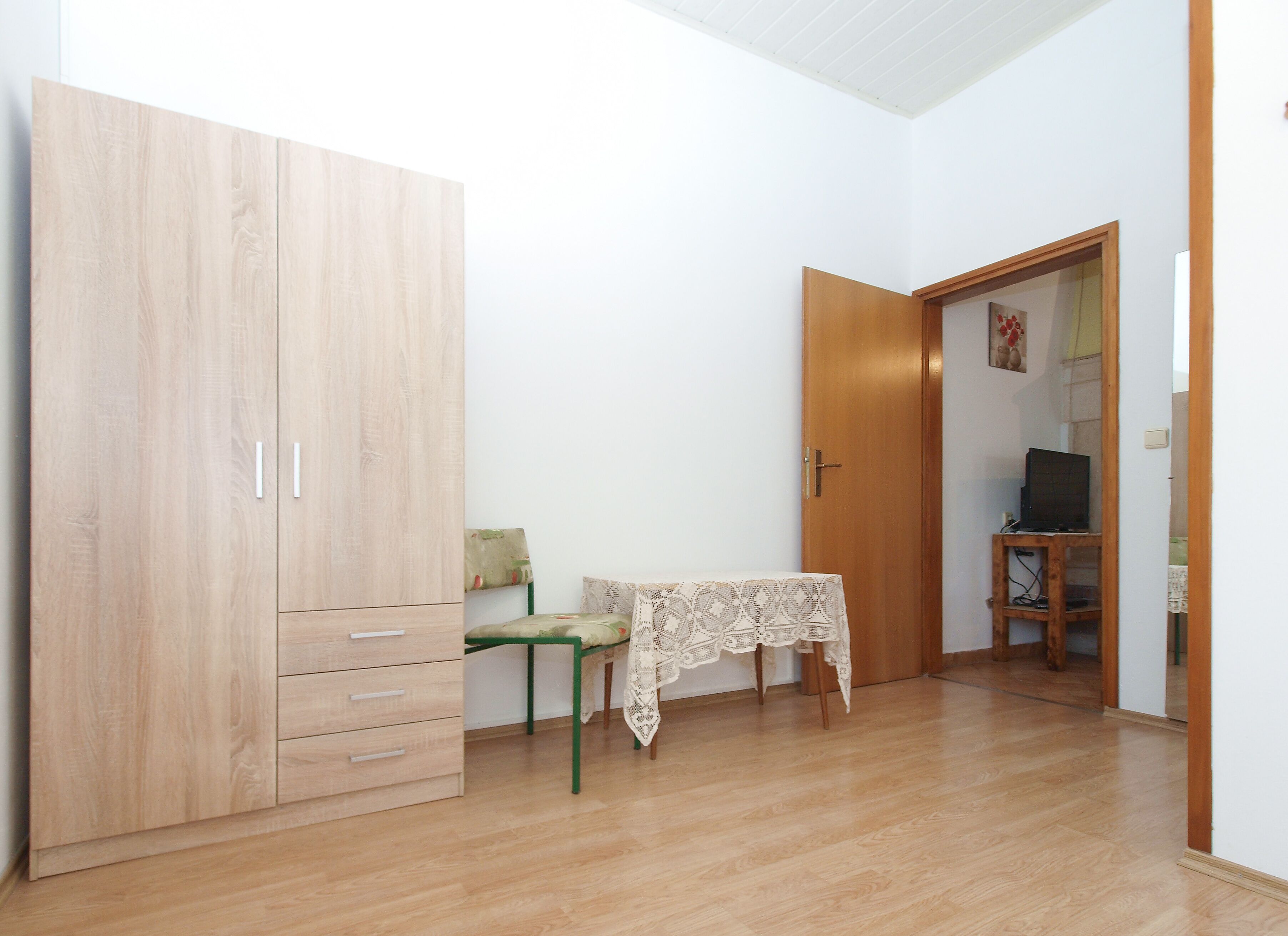 A room with a large wooden wardrobe, a small table with a lace tablecloth, and a television on a wooden stand.