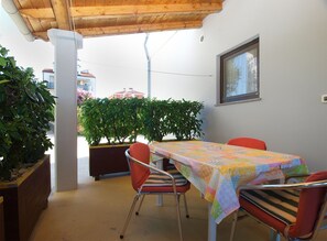 Une terrasse couverte avec une table et des chaises, des plantes en pot et une vue sur les bâtiments en arrière-plan.