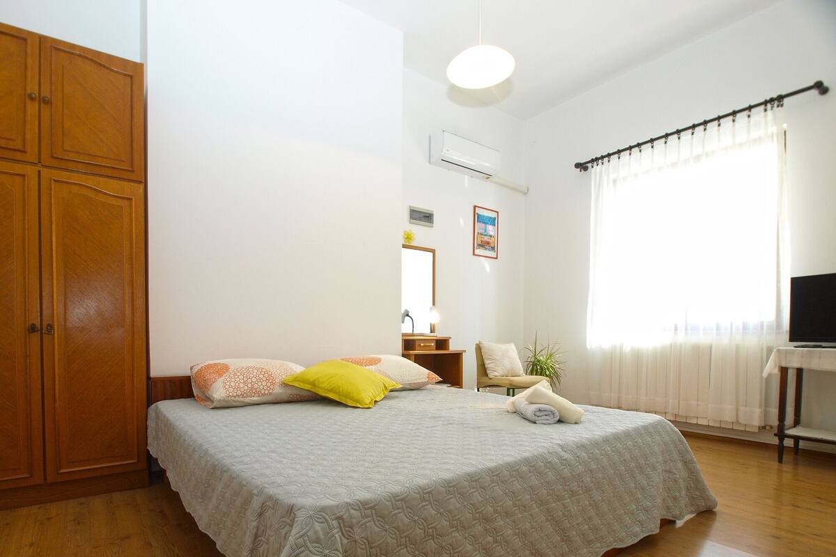 A bedroom with a bed, wooden wardrobe, bedside table, lamp, chair, potted plant, and a TV on a stand.