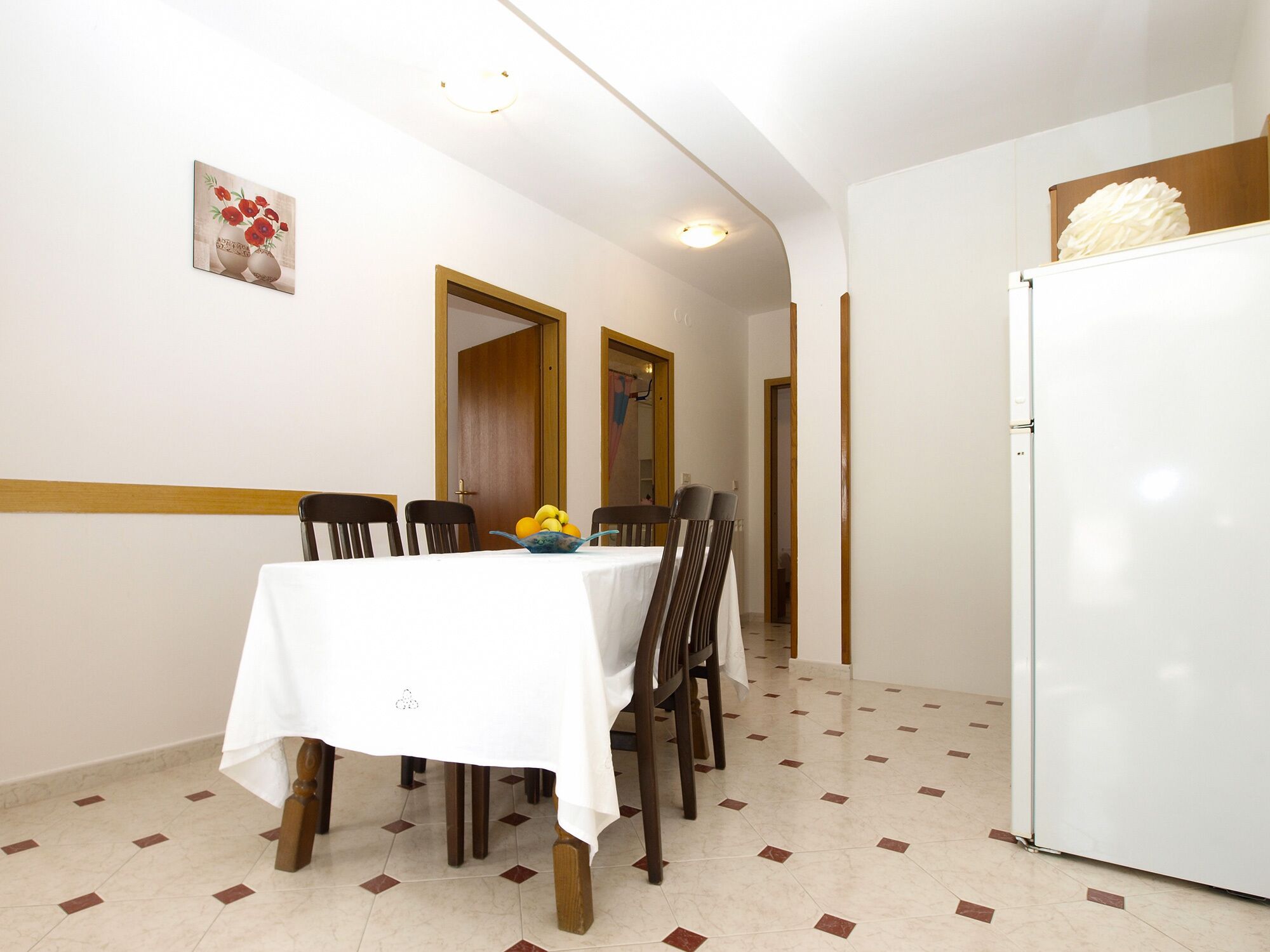 A dining area with a table set for four, a bowl of fruit, and a refrigerator.