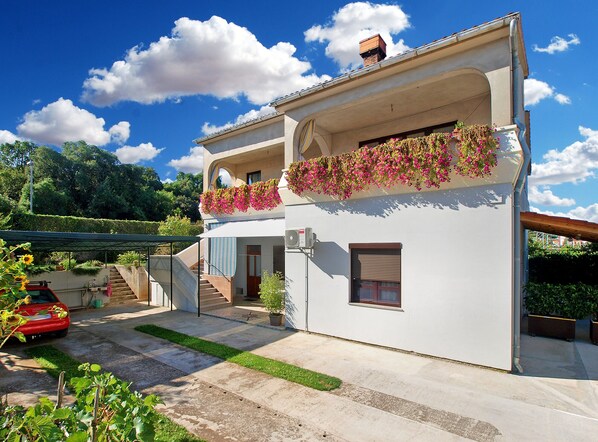 Une maison à deux étages avec un balcon, un escalier et une voiture rouge garée dans l’allée.