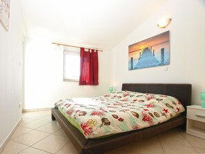 A bedroom with a bed, a window with red curtains, a painting on the wall, and a nightstand with a lamp. - Apartment 1970 (Medulin)