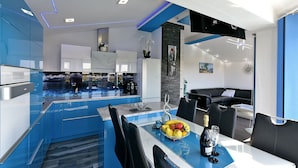 A modern kitchen with blue cabinetry, a dining area with a glass table, and a living area with a black sofa.