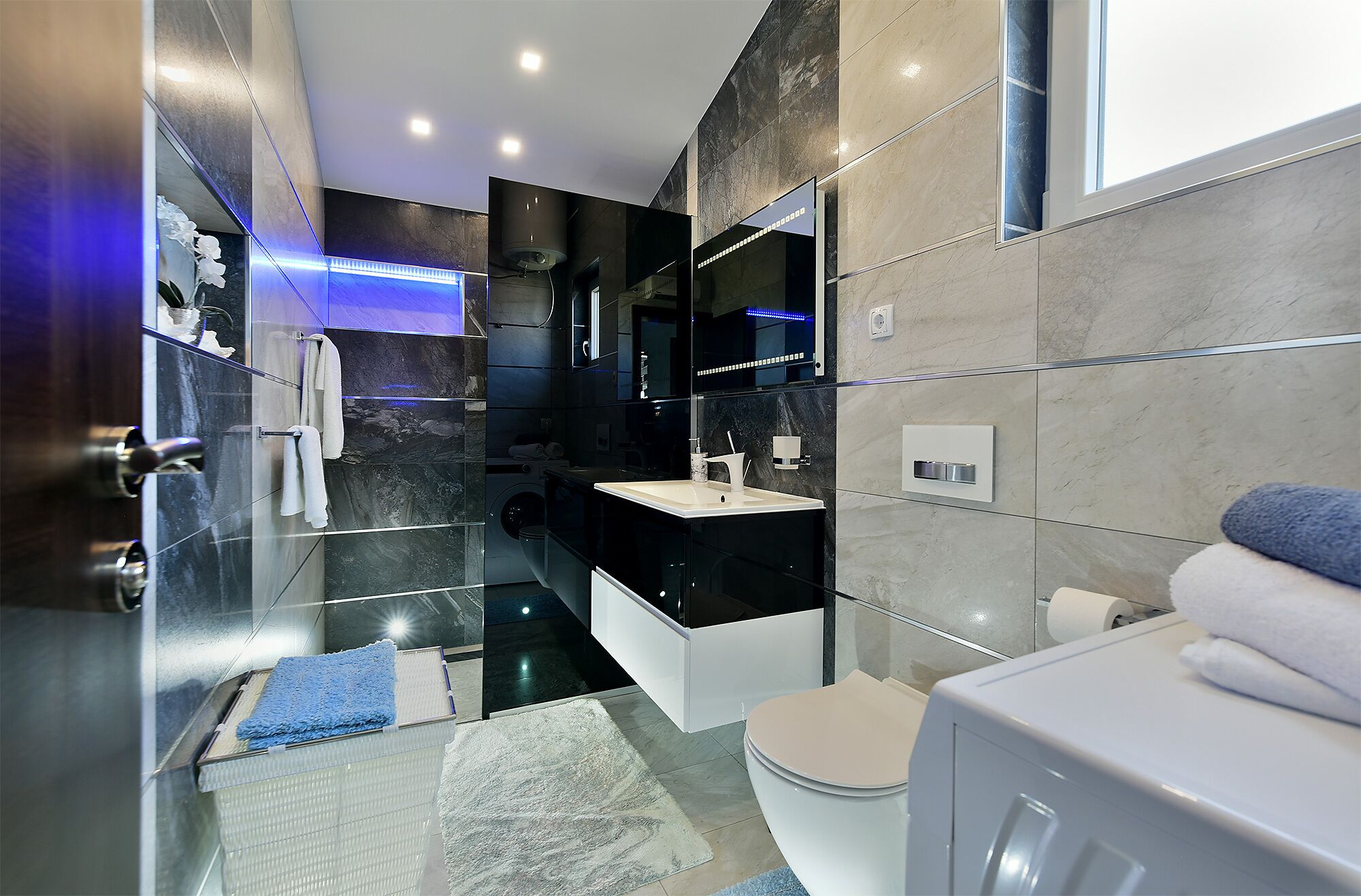 A modern bathroom with a glass shower enclosure, a black and white vanity, a toilet, and a blue rug.