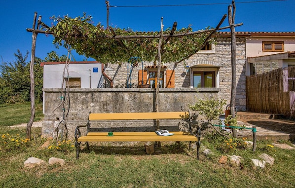 A stone building with a wooden bench in front, a pergola with vines, and a small garden area.
