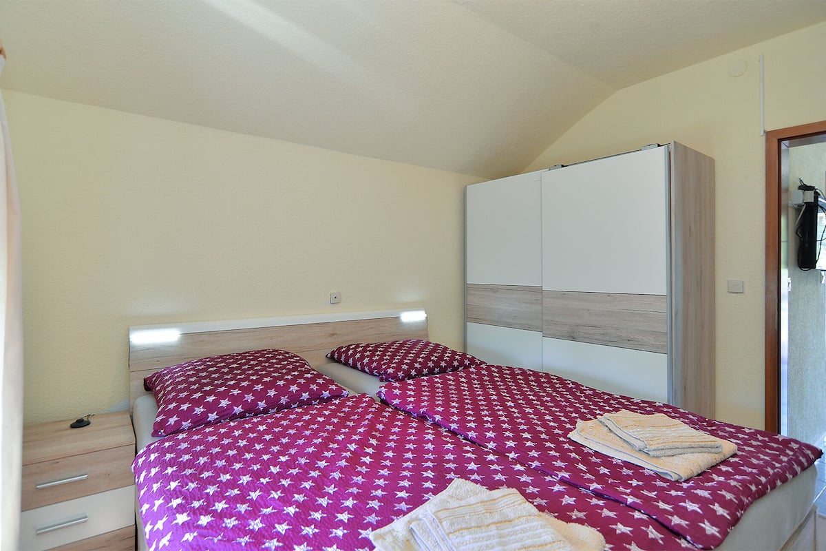 A compact bedroom with two beds, each with a patterned bedspread and pillows, a wardrobe, and a small bedside table.