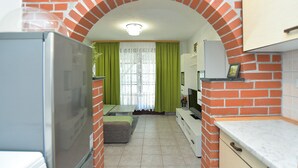 A narrow kitchen with a brick archway, a refrigerator, a television, and a window with green curtains. - Apartment 1976 (Vodnjan)