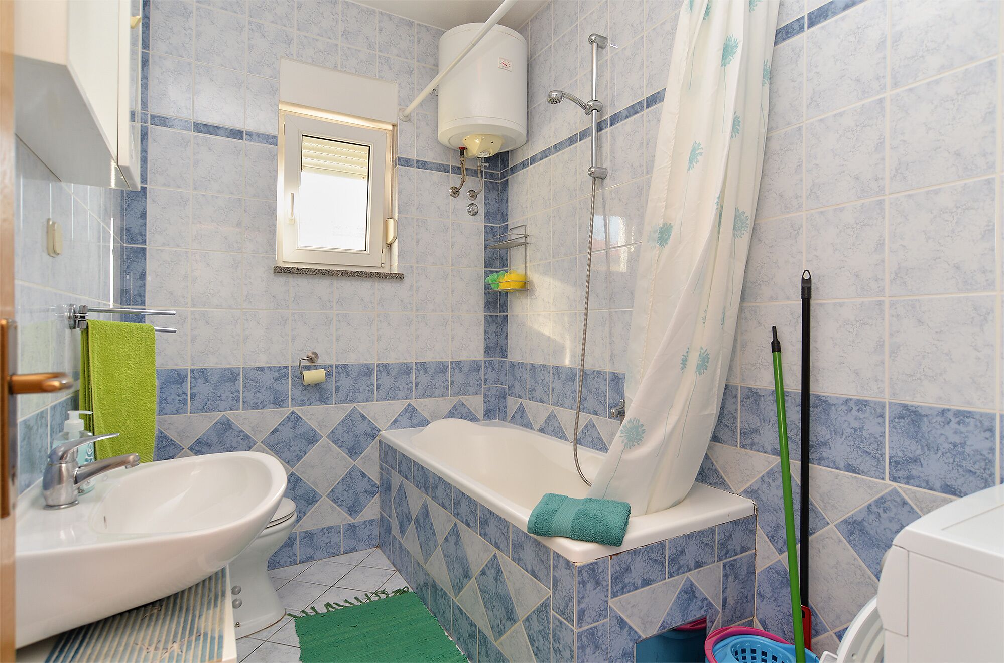 Een badkamer met een bad, een wastafel en een toilet. Er is een douchegordijn, een groene handdoek en schoonmaakmiddelen.