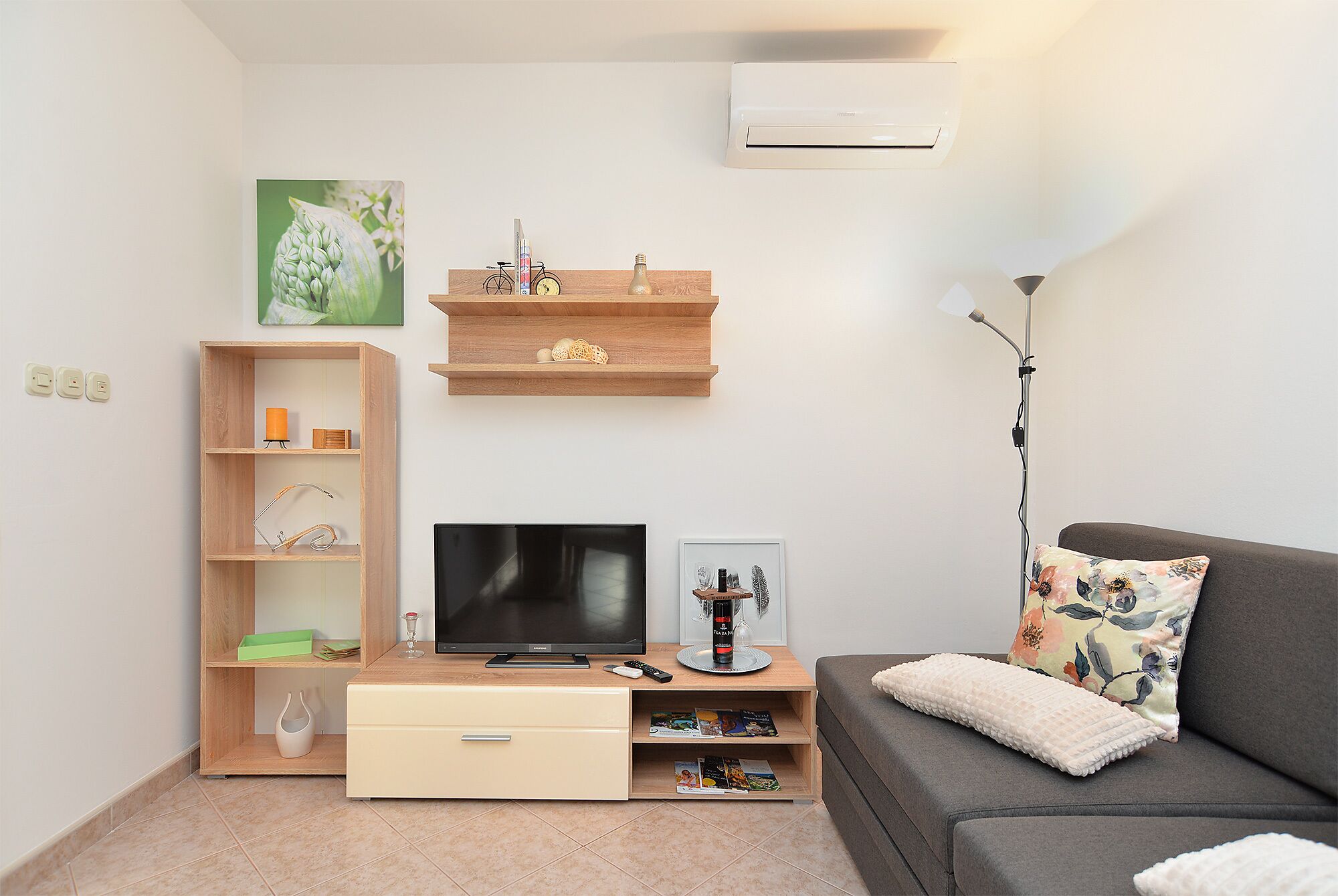 A living room with a flat-screen TV, wooden shelving, a sofa, and a floor lamp.