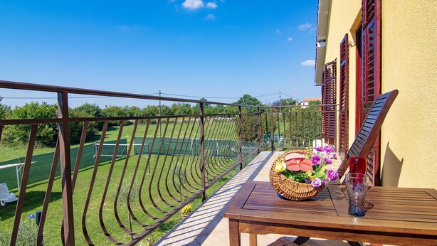 Un balcón con vistas a un campo verde y una cancha de tenis. Hay una mesa con una cesta de mimbre y un vaso.