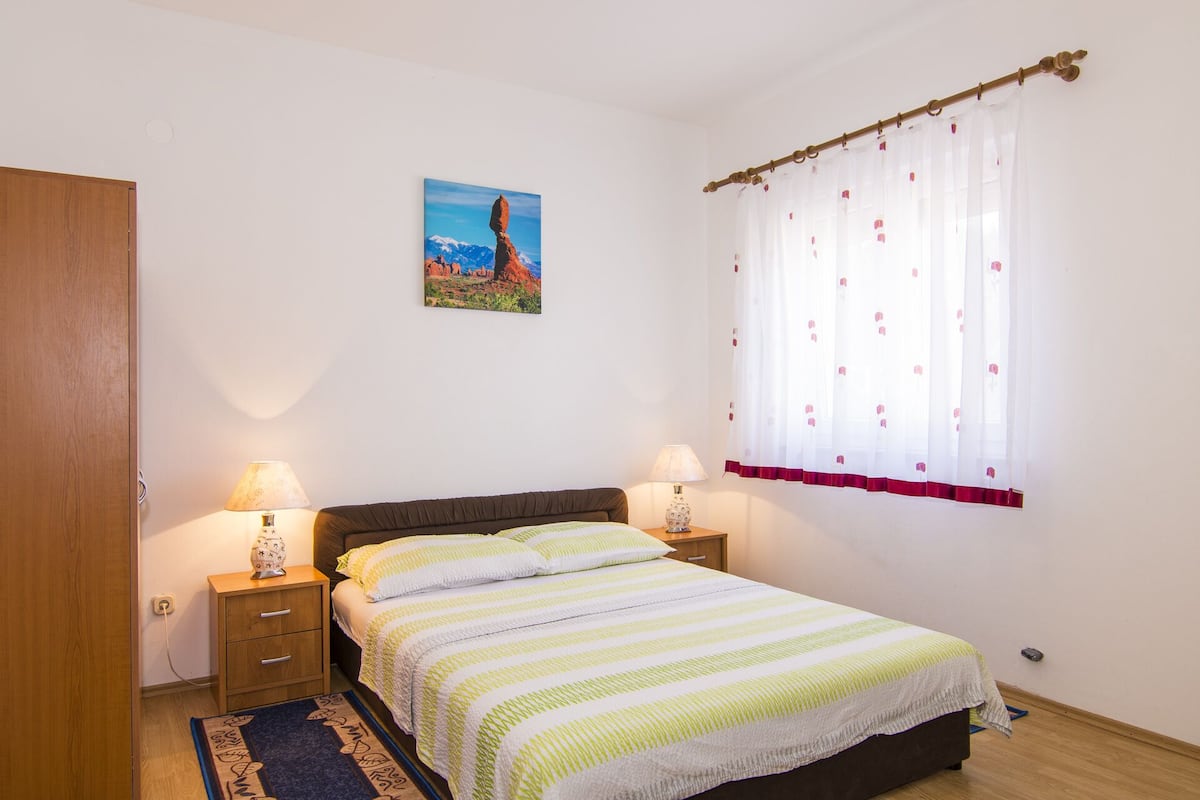 A bedroom with a bed, two bedside tables, a nightstand, a lamp, a window with curtains, and a painting on the wall.