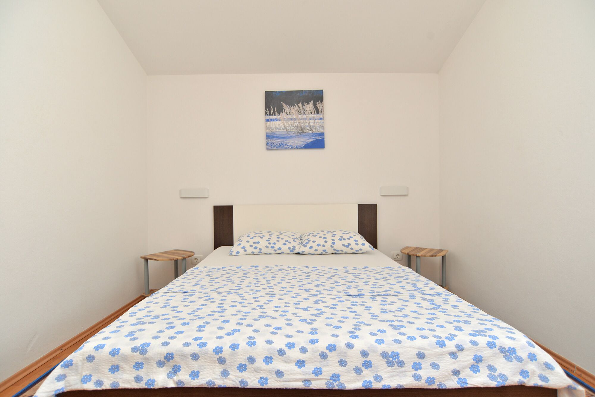 A bedroom with a bed, a nightstand, a wall-mounted light, and a painting above the bed.