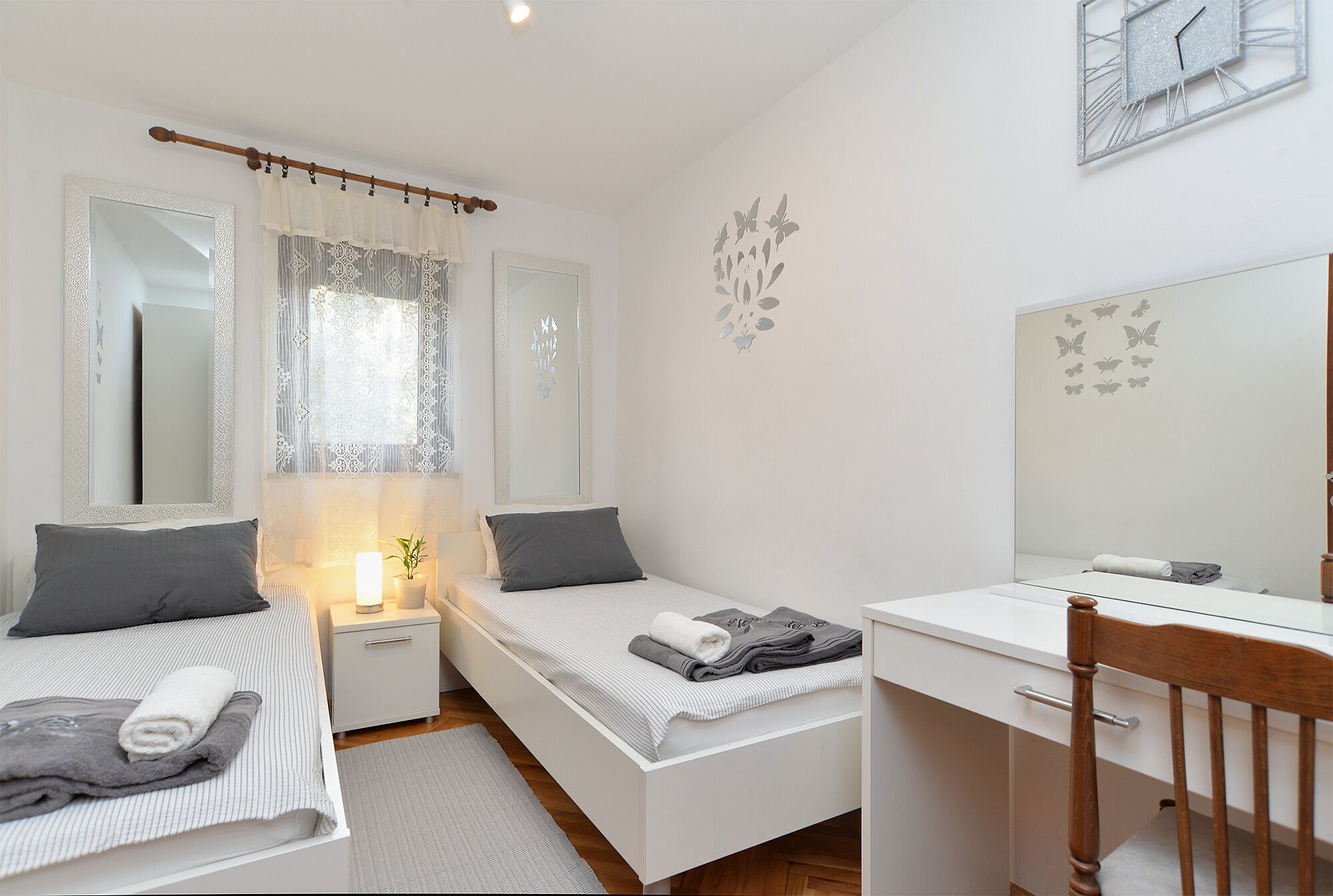 A bedroom with two single beds, a bedside table with a lamp, a desk with a chair, and a large mirror.