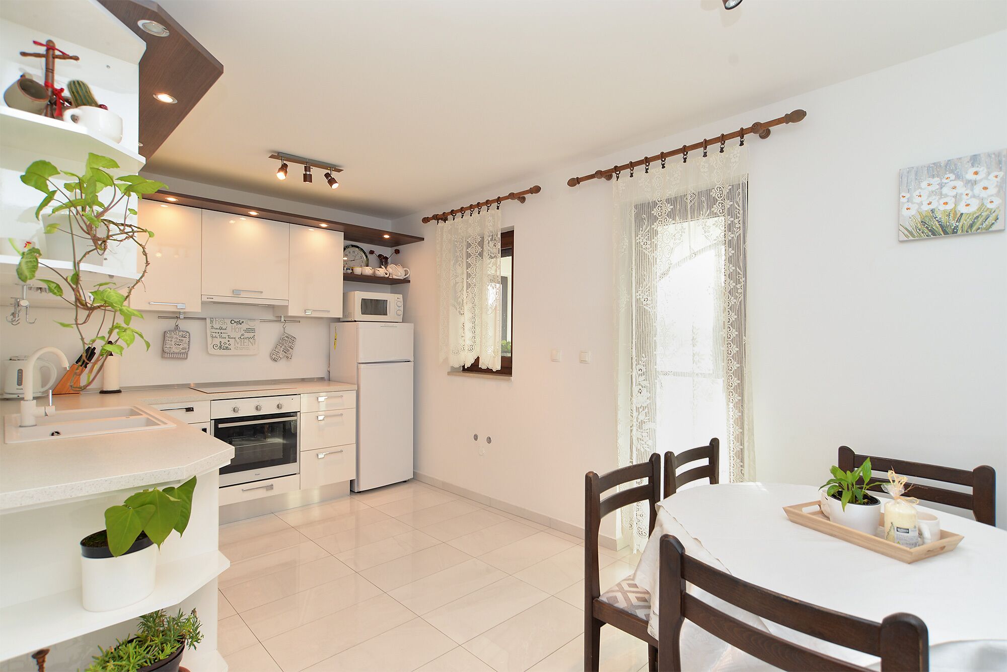 A modern kitchen with white cabinets, a microwave, and a sink. There is a dining table with chairs and a potted plant on it.