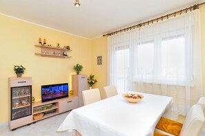 Una sala de estar con televisor, una mesa de comedor con un tazón, y una ventana con cortinas ligeras.