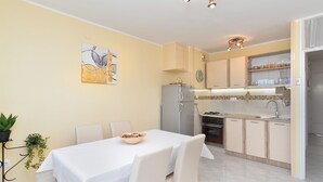 A compact kitchen with a refrigerator, stove, and sink. There is a dining area with a table and chairs. - Apartment 1987 (Pula)