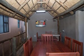 A dining area with a long wooden table and chairs, a television mounted on the wall, and a thatched roof structure. - 848 Guest House (Victoria Falls)