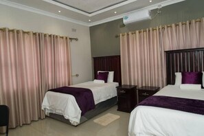 A hotel room with two beds, beige curtains, a dark headboard, and a small bedside table. - 848 Guest House (Victoria Falls)