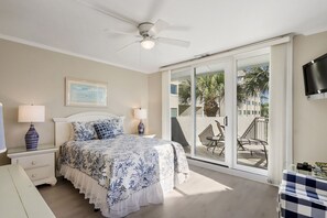 Une chambre à coucher comprenant un lit, des tables de chevet, un ventilateur de plafond et une porte-fenêtre coulissante donnant sur un balcon aménagé avec du mobilier d’extérieur.