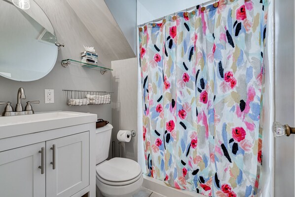 Et badeværelse med en hvid vask, spejl og toilet. Der er et blomstret brusehæng.
