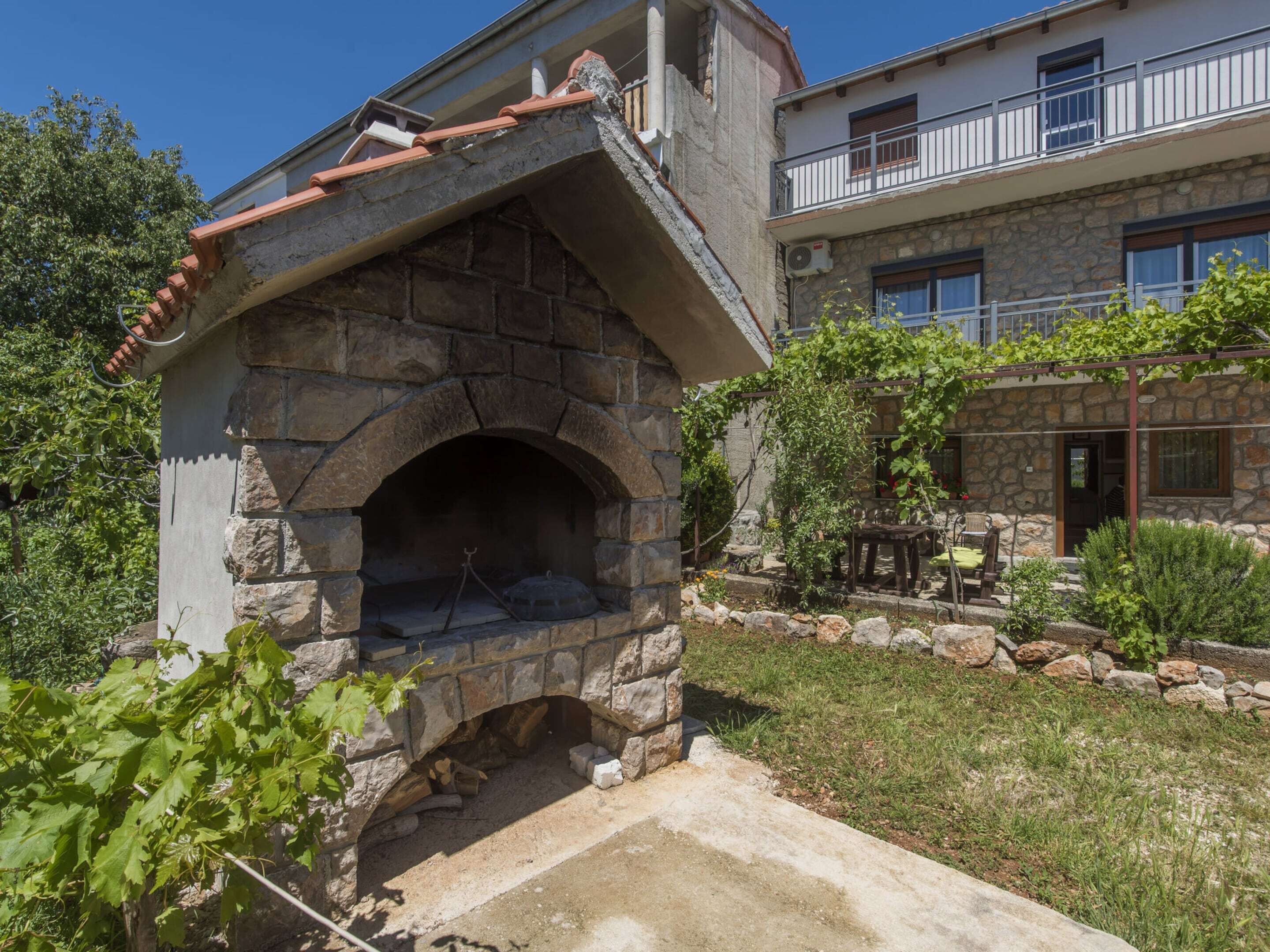 A stone-built outdoor oven with a tiled roof, surrounded by a garden with a table and chairs, and a building with a balcony in the background.