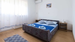 Un dormitorio con cama, dos mesitas de noche, un buró, una ventana con cortinas translúcidas, un equipo de aire acondicionado y una pequeña imagen en la pared.