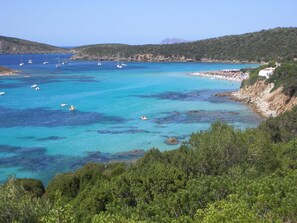 A coastal area with clear turquoise waters, boats, and a beach with people. - Belvilla by OYO Mura Pranu (Sant'Antioco)