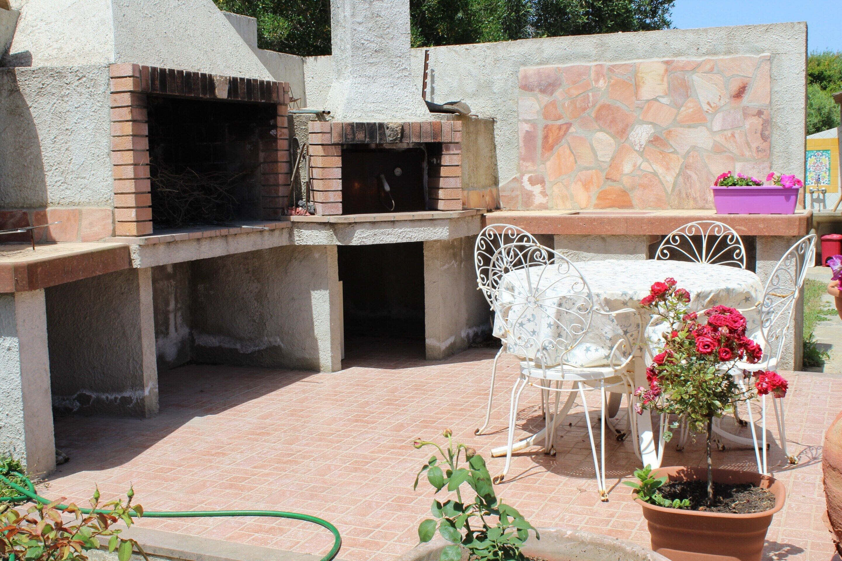 Une terrasse extérieure avec un four en briques, des chaises en métal blanc, une table et un pot de fleurs.