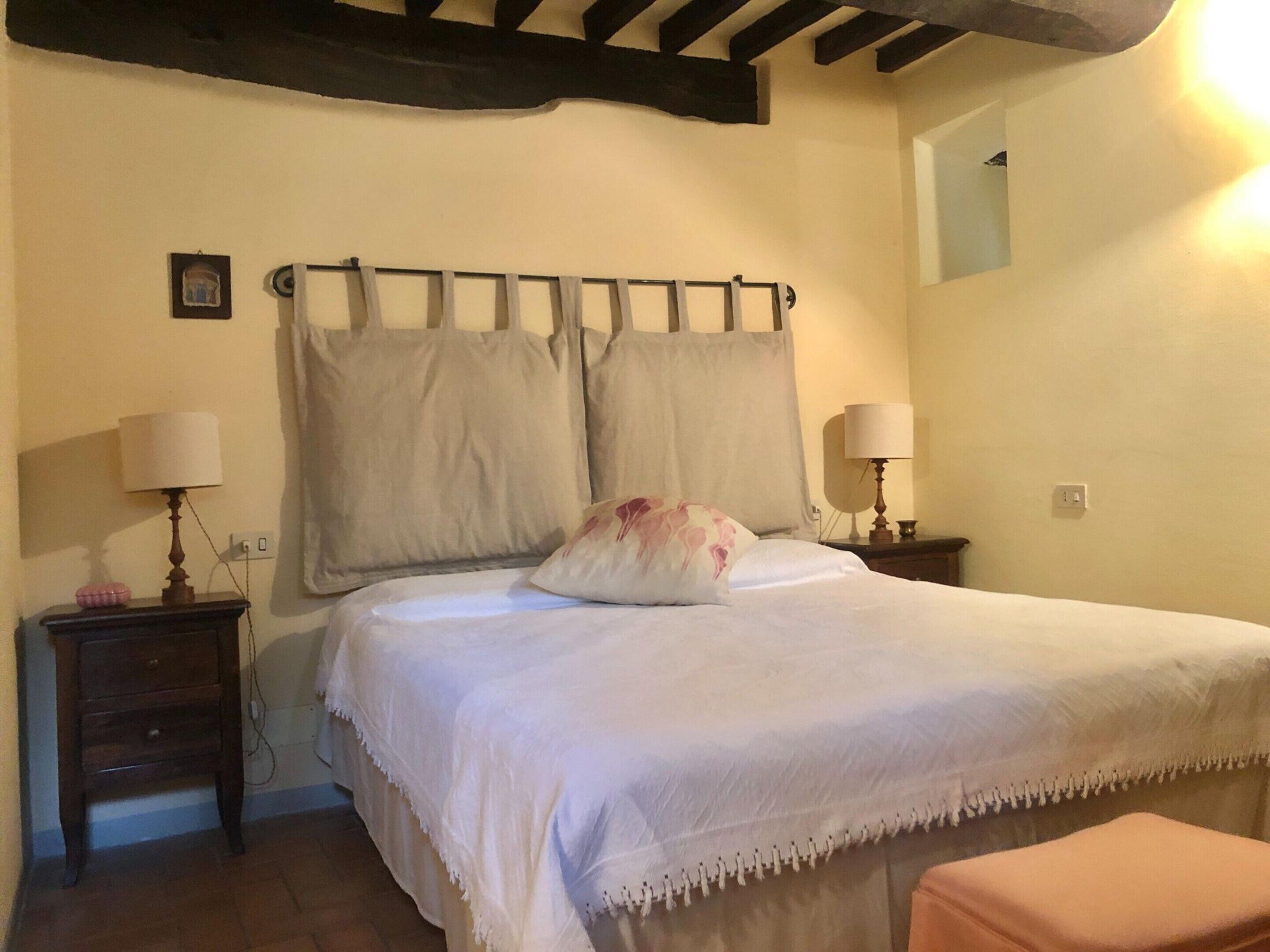 A neatly made bed with a white bedspread and two pillows, a wooden nightstand with a lamp, and a small table with a lamp.