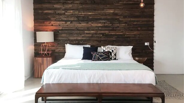 Una camera da letto con testiera in legno, un letto con biancheria da letto bianca e cuscini decorati, una lampada da comodino e un lampadario.