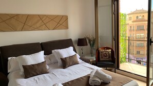 Una camera d'albergo con un letto grande, una finestra vista sui palazzi, una sedia e un tavolo con una lampada.