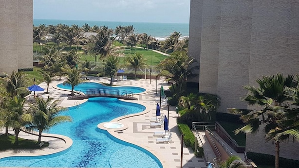 A resort pool area with a curved design, surrounded by palm trees and a view of the ocean. - Scopa Beach Resort Apartments (Aquiraz)