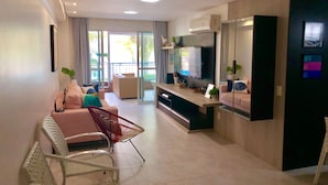 A modern living room with a sofa, chairs, a TV, and a balcony with curtains. - Scopa Beach Resort Apartments (Aquiraz)