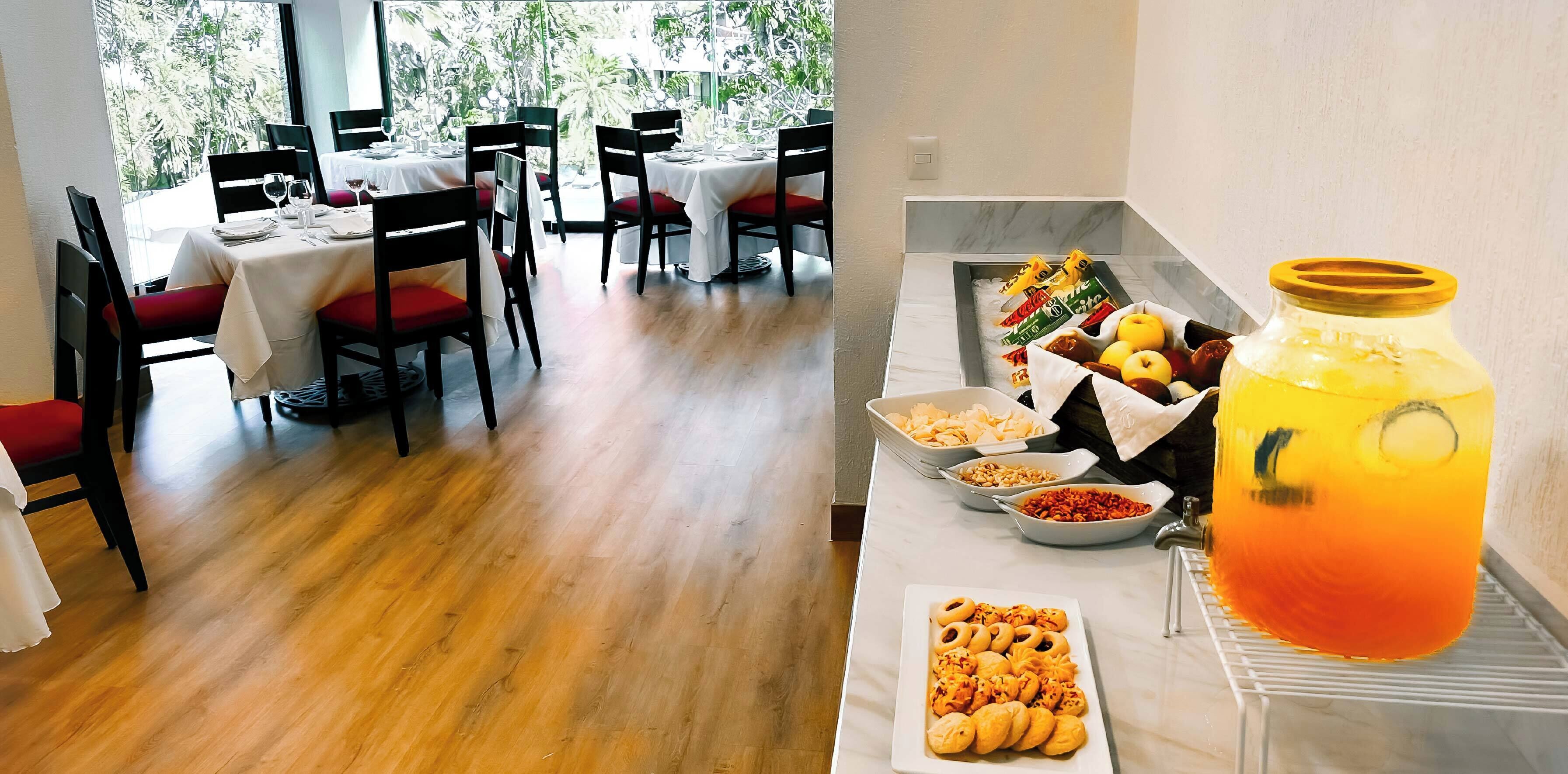 Comedor con mesas dispuestas para cuatro, sillas con cojines rojos y un bufé con diversas opciones de comida y un gran jarro de bebida de naranja.