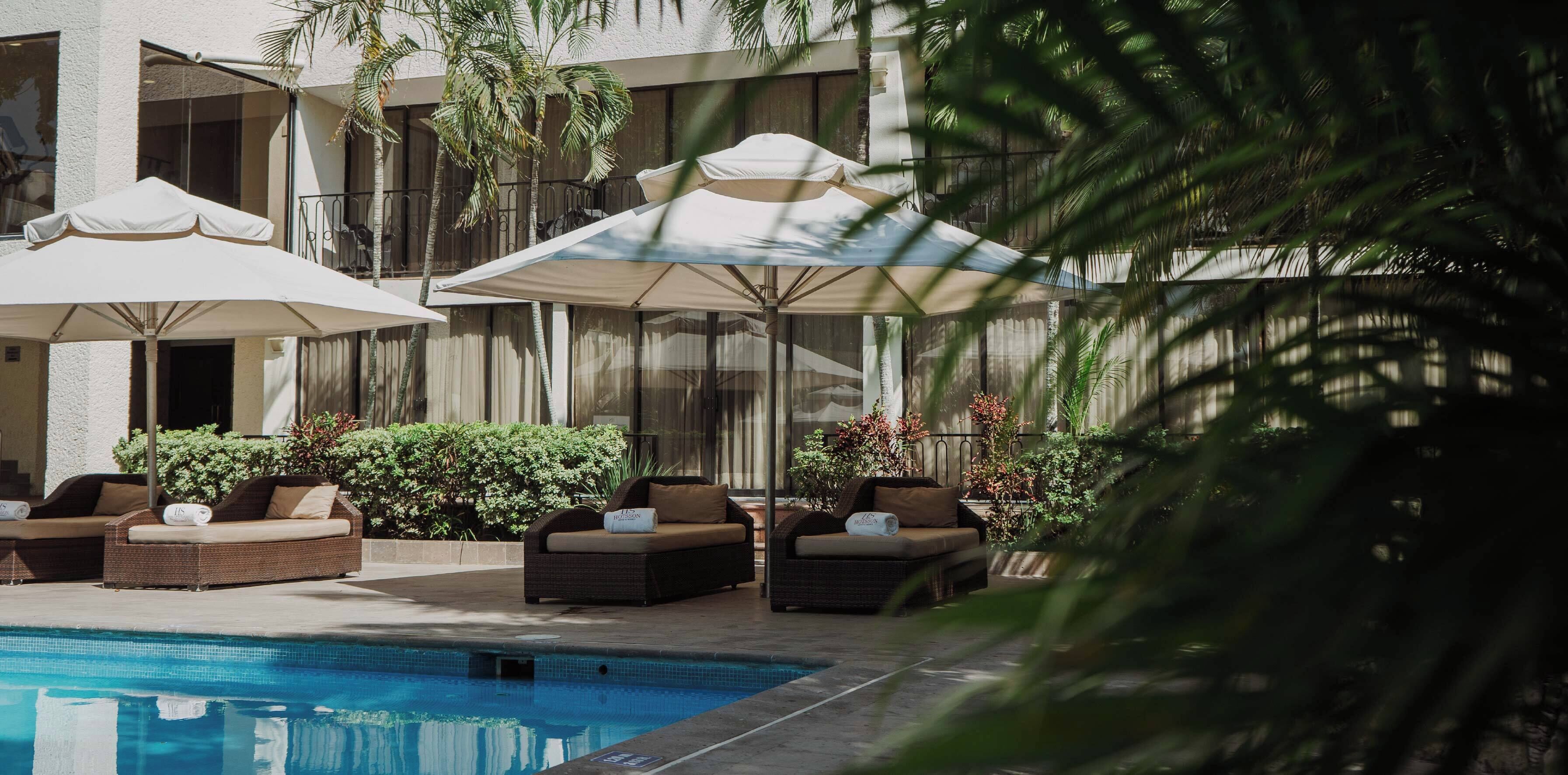 A poolside area with lounge chairs and umbrellas, surrounded by palm trees and a building with large windows.