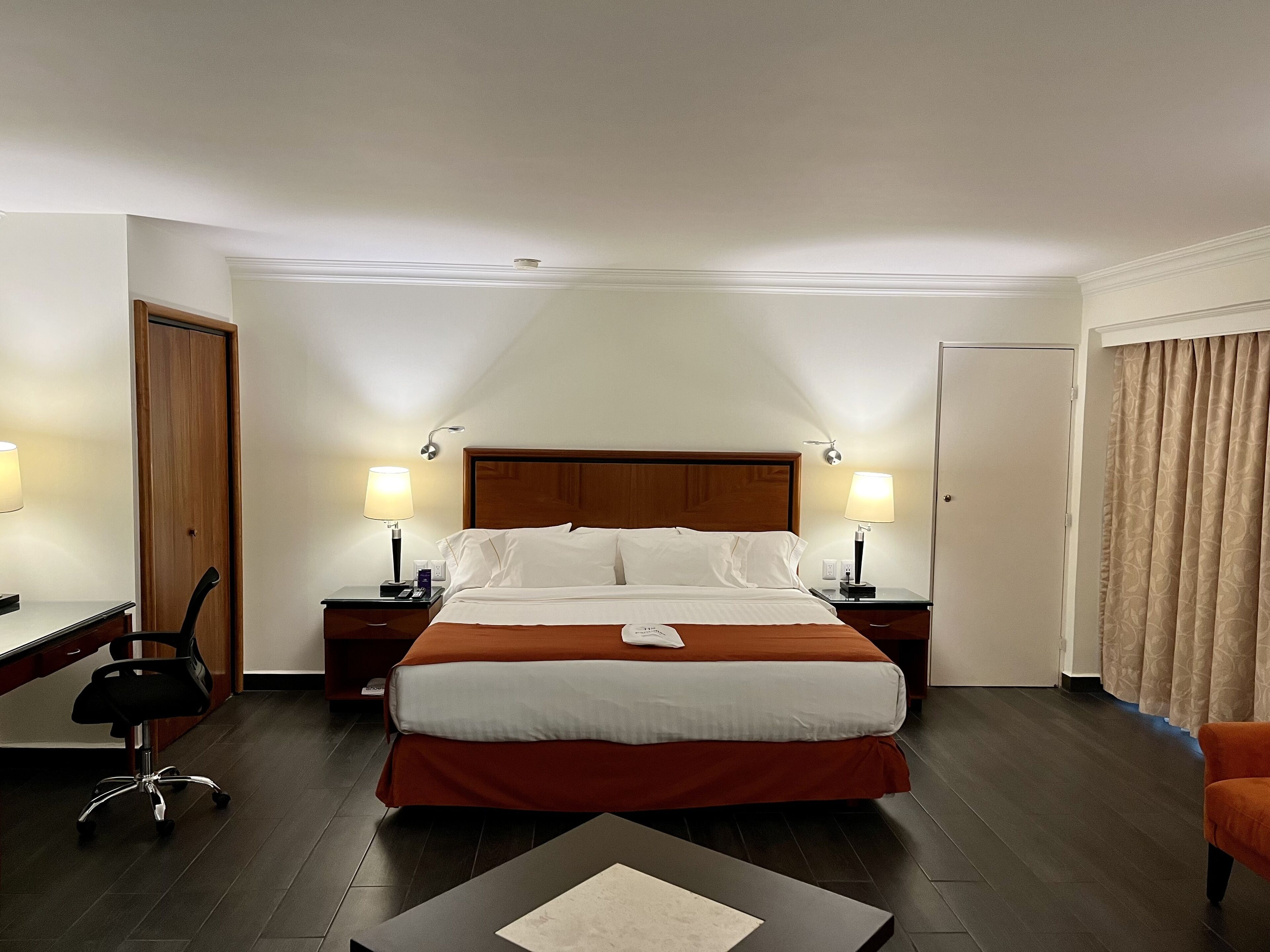 Une chambre d’hôtel avec un grand lit, deux tables de chevet avec des lampes, un bureau avec une chaise et une fenêtre avec des rideaux.