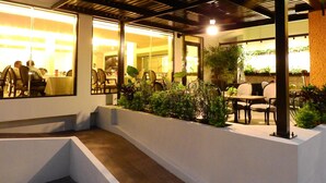 Un espacio de comedor al aire libre con mesas y sillas, rodeado de plantas y una pared blanca.