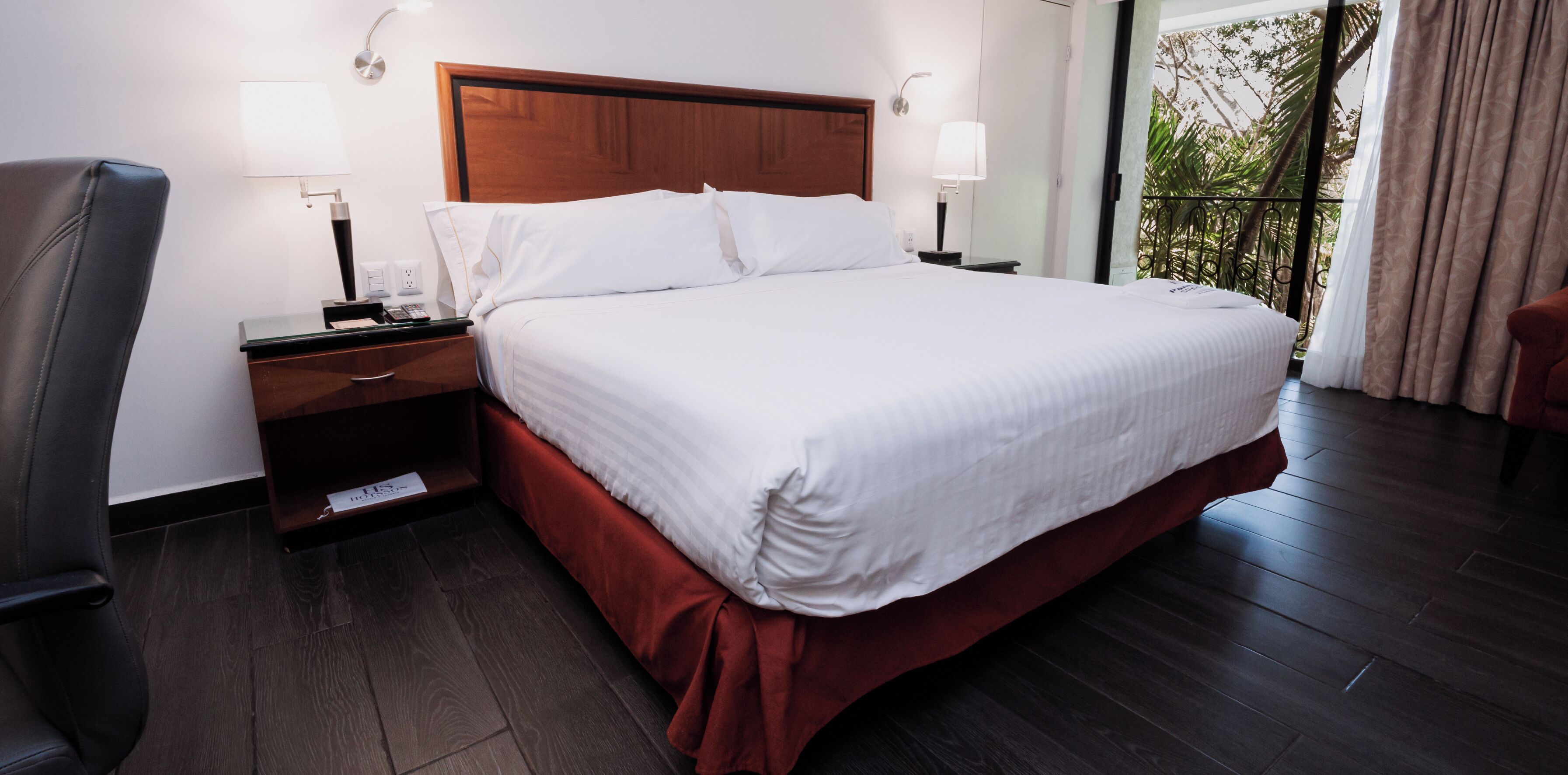 Habitación de hotel con una cama grande, cabecera de madera, dos mesitas de noche con lámparas, una silla y vistas a zonas verdes por la ventana.